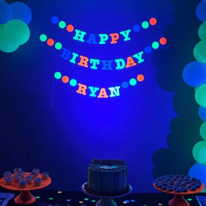 May include: A blue, green, and orange glow-in-the-dark banner that reads "Happy Birthday Ryan". The banner is hanging on a blue wall. There are two cakes on a table in front of the banner. One cake is a layered chocolate cake with white frosting and the other cake is a single-layer cake with blue frosting. There are also cupcakes on the table.