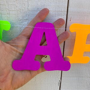May include: A purple letter A, part of a colorful paper banner, is held by a hand against a white wooden background.