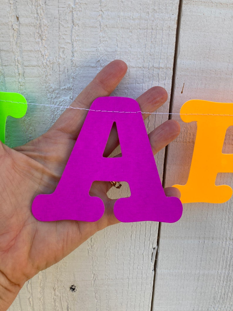 May include: A purple letter A, part of a colorful paper banner, is held by a hand against a white wooden background.