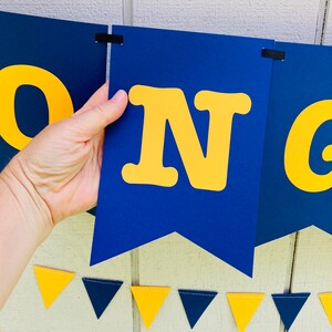 May include: A blue and yellow banner with the word "CONGRADULATIONS" in large yellow letters. The banner is made of cardstock and is strung together with black ribbon. There are also small blue and yellow triangle flags strung below the banner.