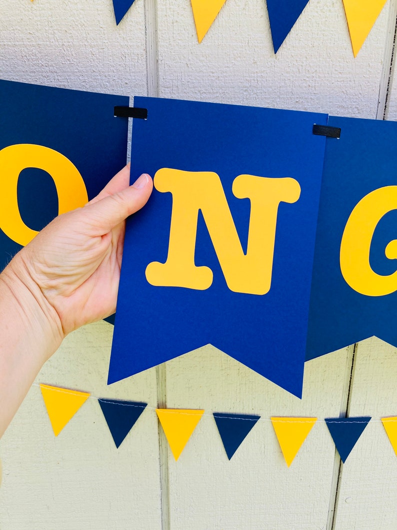 May include: A blue and yellow banner with the word "CONGRADULATIONS" in large yellow letters. The banner is made of cardstock and is strung together with black ribbon. There are also small blue and yellow triangle flags strung below the banner.