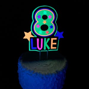 Puede incluir: Un adorno para pastel verde que brilla en la oscuridad con el n&uacute;mero 8 y el nombre "LUKE" en colores ne&oacute;n. El n&uacute;mero 8 tiene estrellas de colores en su interior.