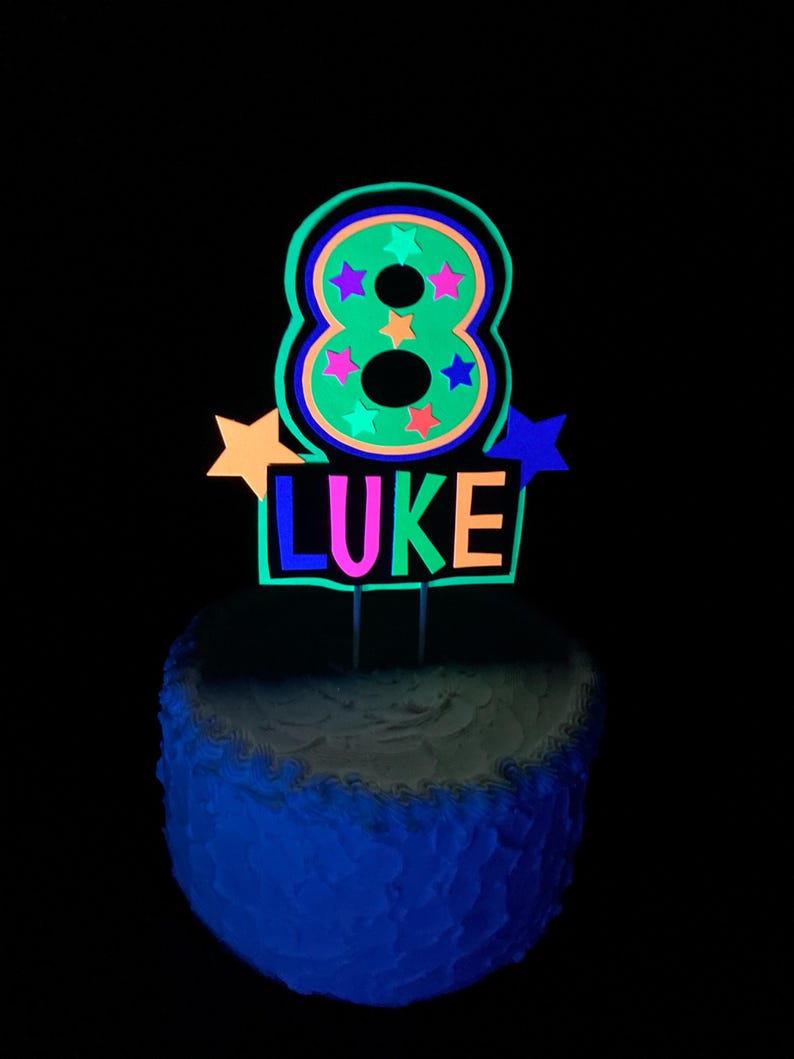 Puede incluir: Un adorno para pastel verde que brilla en la oscuridad con el n&uacute;mero 8 y el nombre "LUKE" en colores ne&oacute;n. El n&uacute;mero 8 tiene estrellas de colores en su interior.