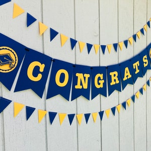 May include: A blue and gold graduation banner with the word "CONGRATS" and a graduation cap icon. The banner is strung with a black string and is decorated with blue and gold triangle pennants.