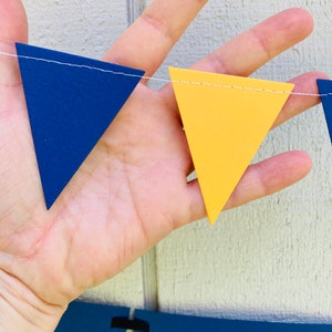 May include: A string of blue and yellow triangle pennants, strung together with white thread. The pennants are made of felt and have a stitched edge.