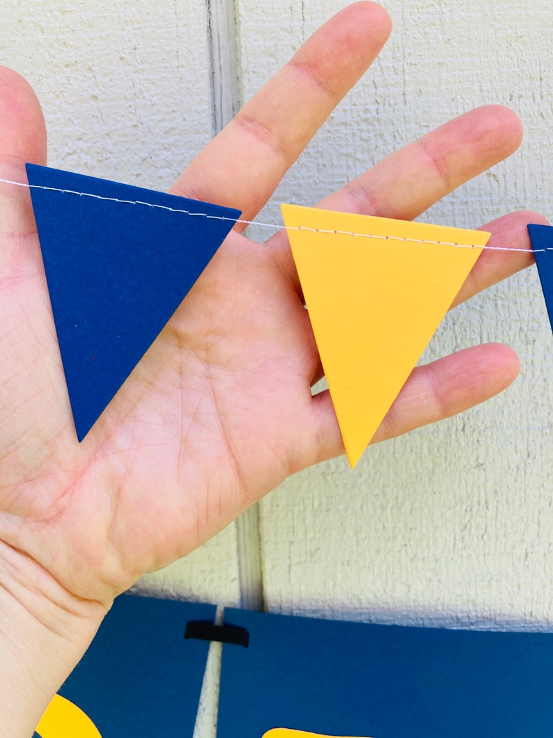 May include: A string of blue and yellow triangle pennants, strung together with white thread. The pennants are made of felt and have a stitched edge.