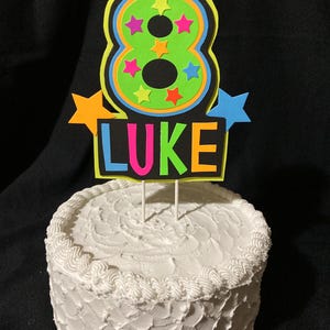 Puede incluir: Un adorno para pastel de color verde ne&oacute;n y negro con un gran n&uacute;mero 8, rodeado de estrellas de colores, y el nombre "LUKE" escrito debajo.