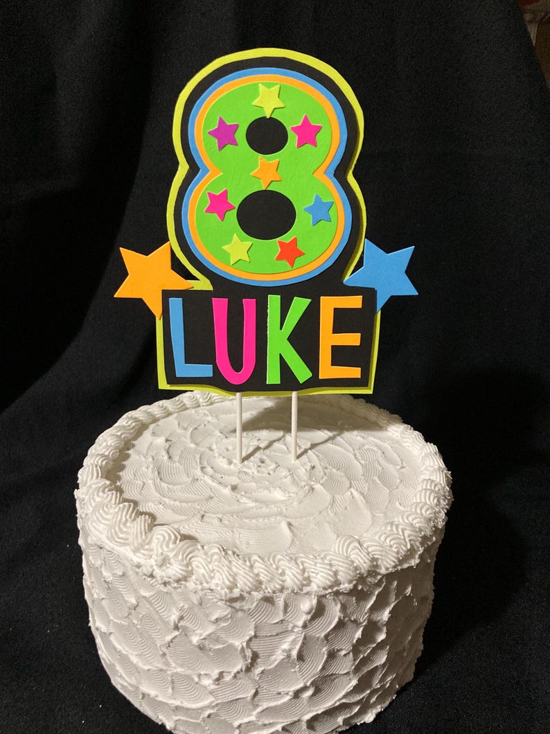 Puede incluir: Un adorno para pastel de color verde ne&oacute;n y negro con un gran n&uacute;mero 8, rodeado de estrellas de colores, y el nombre "LUKE" escrito debajo.
