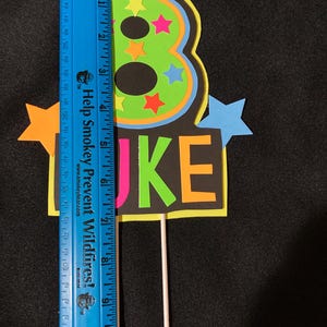 Puede incluir: Un n&uacute;mero ocho verde ne&oacute;n y amarillo con estrellas negras y de colores, un fondo negro con la palabra "LUKE" en letras de colores, y un palo blanco unido a la parte inferior.