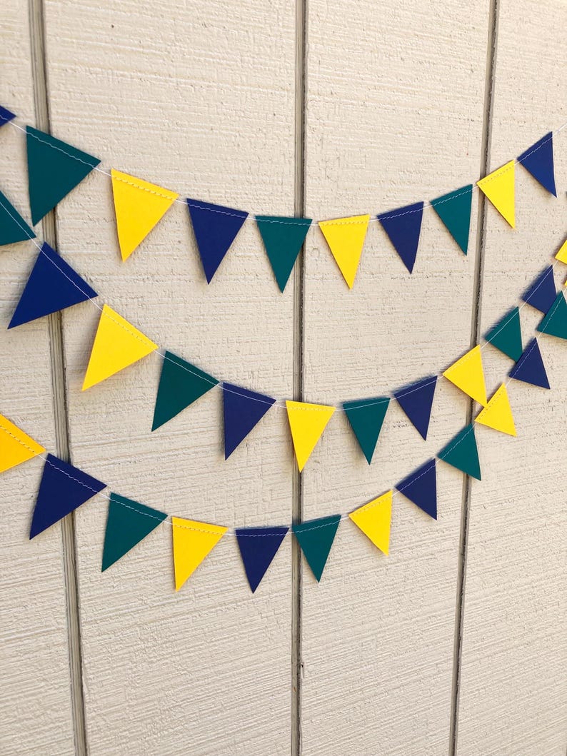 Peut inclure: Trois guirlandes de fanions triangulaires color&eacute;s jaunes, turquoise et bleu marine sont suspendues sur un fond en bois blanc. Les fanions sont espac&eacute;s uniform&eacute;ment et reli&eacute;s par un fil blanc, cr&eacute;ant une d&eacute;coration festive.