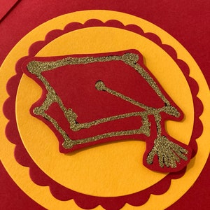 May include: A red and gold graduation cap with glitter accents on a red and yellow card.