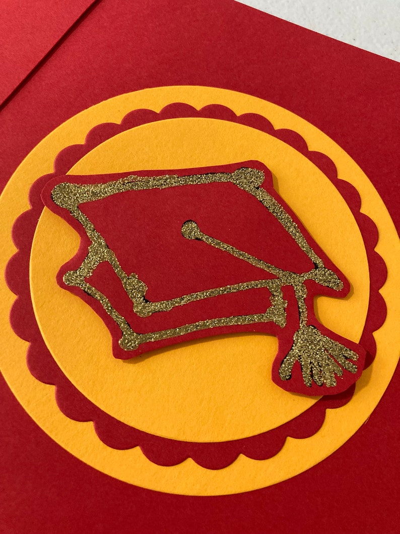 May include: A red and gold graduation cap with glitter accents on a red and yellow card.
