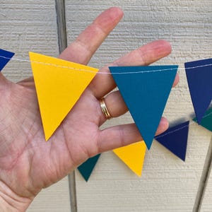 Peut inclure: Une guirlande color&eacute;e de drapeaux triangulaires jaunes, bleu sarcelle et bleu marine. Les drapeaux sont enfil&eacute;s sur une ficelle blanche, cr&eacute;ant une d&eacute;coration festive. La guirlande est tenue par une main.