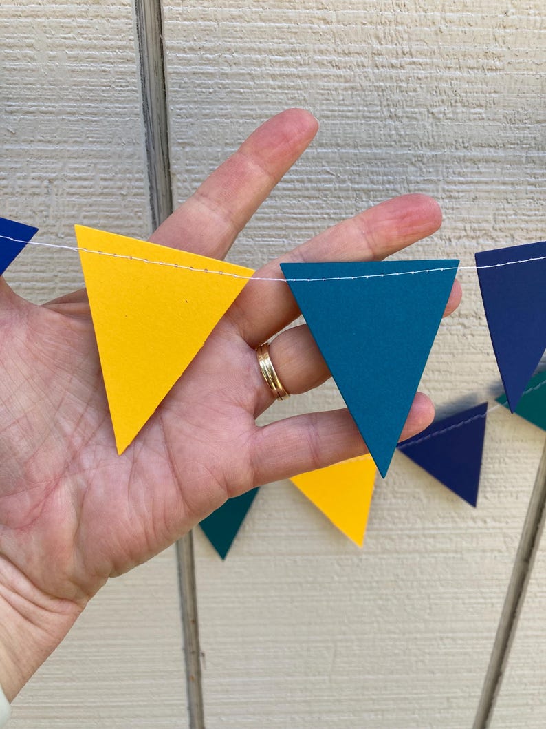 Peut inclure: Une guirlande color&eacute;e de drapeaux triangulaires jaunes, bleu sarcelle et bleu marine. Les drapeaux sont enfil&eacute;s sur une ficelle blanche, cr&eacute;ant une d&eacute;coration festive. La guirlande est tenue par une main.