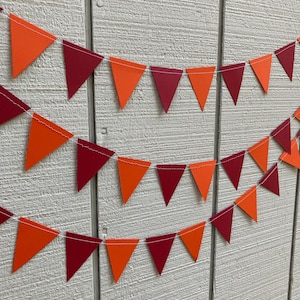 May include: A festive banner composed of orange and burgundy triangular flags strung on white string. The pennants are arranged in three rows against a white wooden background, creating a decorative display.