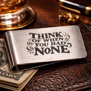 Könnte beinhalten: Ein silberner Geldclip mit dem eingravierten Text "THINK OF WHEN YOU HAD NONE." Der Clip liegt auf einem braunen Lederportemonnaie mit einem Stapel US-Dollar-Scheine. Im Hintergrund ein Glas mit bernsteinfarbener Flüssigkeit.