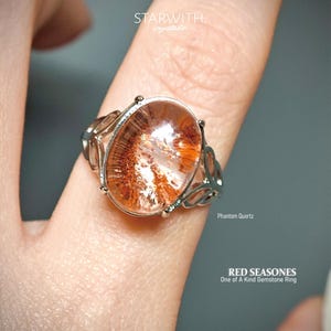 May include: A silver ring featuring a large, oval-shaped phantom quartz gemstone with red and orange inclusions. The ring has an ornate, openwork design on the band. Text on the image reads "STARWITH crystals", "Phantom Quartz", and "RED SEASONES One of A Kind Gemstone Ring".