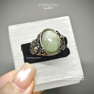 May include: An adjustable ring featuring an oval green phantom quartz gemstone set in a silver-toned metal band with a textured, web-like pattern. Text on the image reads STARWITH crystals, Green Phantom Quartz, and GREEN SPIDER One of A Kind Gemstone Adjustable Rings.
