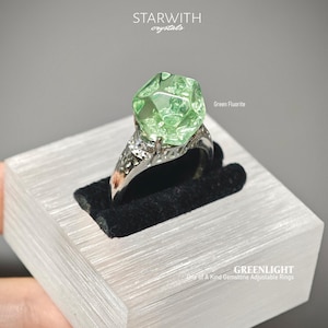 May include: An adjustable ring featuring a faceted, light green fluorite gemstone set in a silver-toned metal band with intricate cut-out details. The text STARWITH crystals, Green Fluorite, and GREENLIGHT One of A Kind Gemstone Adjustable Rings is visible.