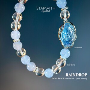 May include: A blue aquamarine gemstone wrapped in wire is the centerpiece of this bracelet. The bracelet is made with clear quartz and light blue beads. The bracelet is labeled "Raindrop Stress Relief & Inner Peace Crystal Jewelry".