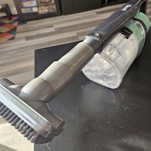 May include: A Shark brand handheld vacuum cleaner. The vacuum has a black handle and a gray brush head. The dustbin is clear and green. The vacuum is on a black surface.