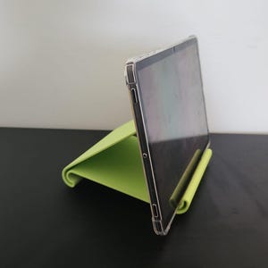 Puede incluir: Una tableta apoyada en un soporte verde lima. La tableta está en una funda transparente. El soporte está hecho de un material plegado y curvado. El fondo es una pared blanca y una superficie negra.