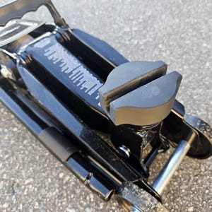 Puede incluir: Un gato hidráulico negro con un sillín de goma. El gato tiene un asa y ruedas para facilitar el movimiento. El gato está diseñado para levantar vehículos y otros objetos pesados.