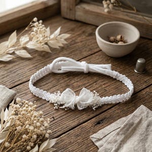 Könnte beinhalten: Ein weißes Stirnband mit zarter Spitzenborte und einer zentralen Schleife, die mit kleinen getrockneten weißen Blumen verziert ist. Das Band besteht aus weichem Stoff mit einer Bindeschleife am Rücken und zeigt ein detailliertes Webmuster.