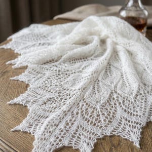Peut inclure: Un châle triangulaire blanc présentant un motif de dentelle ajourée complexe avec des détails en forme de feuilles et un bord festonné pointu. Le tissu en maille délicat et semi-transparent est drapé pour mettre en valeur sa texture.