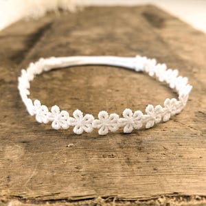 Pode incluir: Uma faixa de cabelo floral branca com um delicado design de margaridas. A faixa é feita de um material macio e elástico, perfeita para adicionar um toque de elegância a qualquer penteado. Apresentada em uma superfície de madeira rústica.
