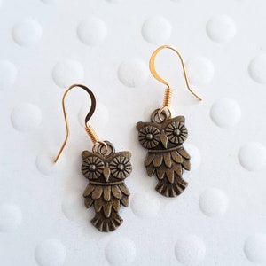 May include: A pair of gold dangle earrings with bronze owl charms. The owls have detailed features, including eyes, beaks, and feathers.