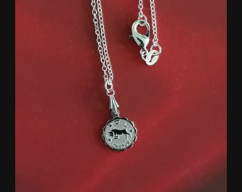May include: Silver necklace with a silver pendant featuring the Taurus zodiac sign. The pendant is round and has a black bull design with the word "Taurus" inscribed on it.