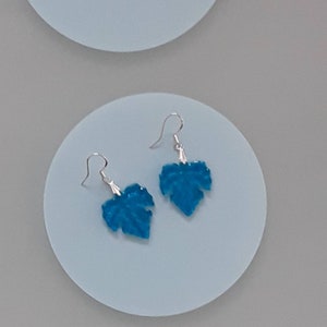 May include: A pair of silver earrings with blue leaf-shaped charms. The leaves are a vibrant turquoise color and have a textured surface.
