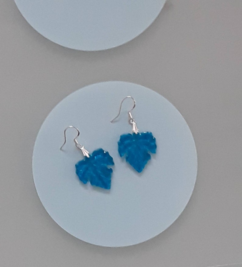 May include: A pair of silver earrings with blue leaf-shaped charms. The leaves are a vibrant turquoise color and have a textured surface.
