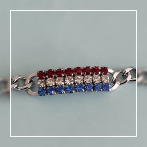 May include: A silver chain bracelet with a red, white, and blue rhinestone charm. The charm is in the shape of a rectangle and has a row of red rhinestones, a row of white rhinestones, and a row of blue rhinestones.