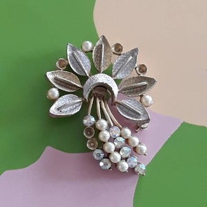 May include: Silver-toned vintage brooch with a floral design. The brooch features stylized leaves, faux pearls, and dangling crystal accents. The leaves have a textured finish, and the brooch has a classic, elegant appearance.