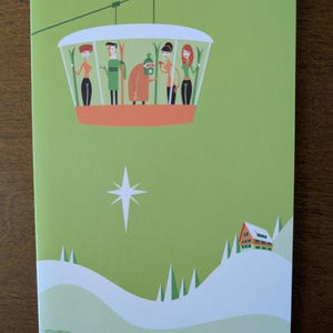 May include: A green greeting card with a ski lift illustration. The card features a ski lift with five people inside, set against a light green background. A white star and a small house on a snowy hill are also depicted.