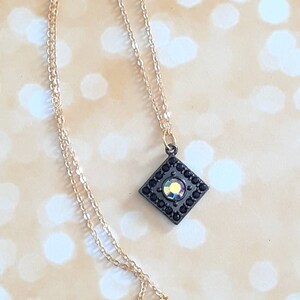 May include: A gold-colored chain necklace with a square pendant. The pendant is black with a central iridescent stone and is framed by small black stones. The necklace is displayed on a light, bokeh-style background.