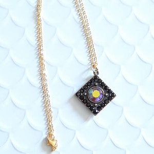 May include: A gold chain necklace with a square pendant featuring a black border and a multi-colored iridescent stone in the center.