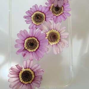 May include: Clear phone case with embedded dried pink and yellow flowers. The flowers are arranged diagonally across the case, showcasing their delicate petals and intricate details. The case provides a stylish and protective design.