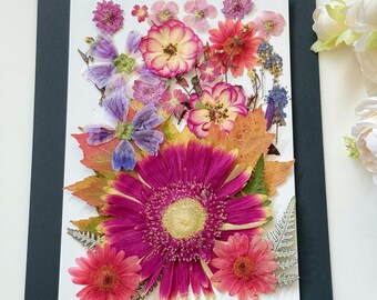 Op de afbeelding: Een kunstwerk van geperste bloemen met een roze gerbera, roze en paarse bloemen en herfstbladeren. Het kunstwerk is ingelijst met een zwarte passe-partout en heeft het woord "Dahlia" onderaan.