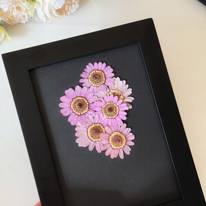 May include: A framed floral art piece featuring a cluster of pink and yellow dried flowers. The flowers are arranged on a black background within a black rectangular frame. The artwork is a close-up shot, highlighting the delicate details of the petals.