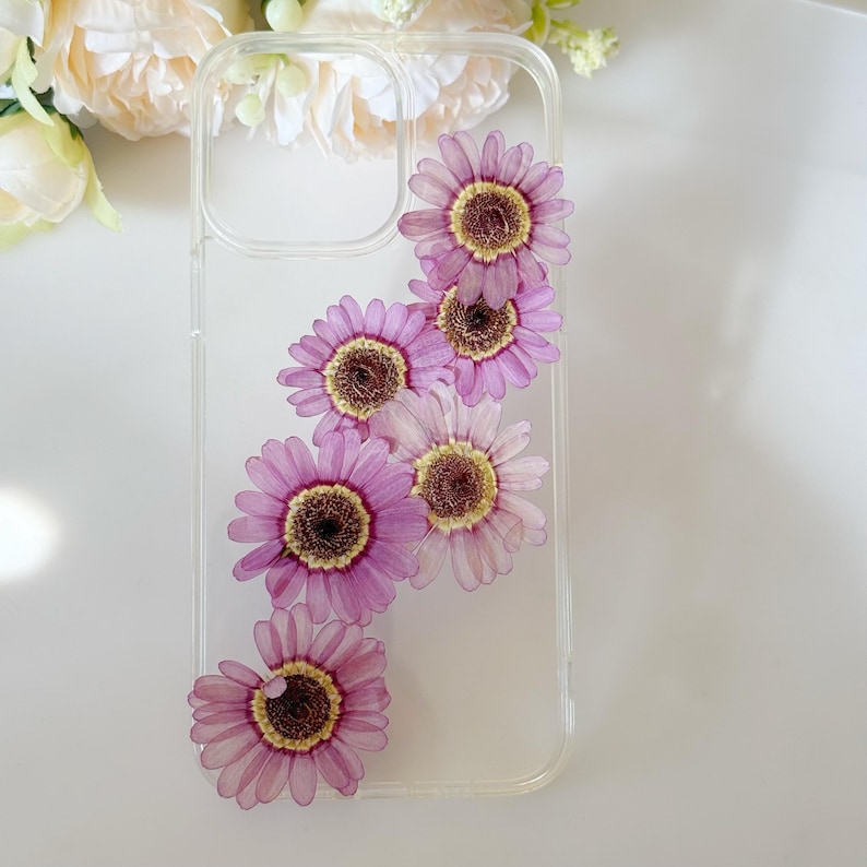 May include: Clear phone case adorned with pressed purple and yellow daisies. The flowers are arranged diagonally across the case, creating a delicate and natural aesthetic. The case is made of clear plastic.
