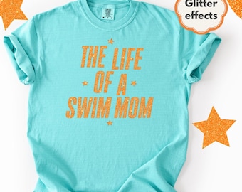 May include: A turquoise t-shirt with the text "THE LIFE OF A SWIM MOM" in orange glitter lettering. The shirt has a crew neck and short sleeves. Orange glitter star accents are above and below the text. The words "Glitter effects" are in a cloud-shaped graphic.