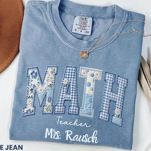 May include: A blue jean colored t-shirt featuring the word MATH in large block letters with varied blue and white floral and gingham patterns. Below the letters, the text reads Teacher Mrs. Rausch in white script. A gold necklace rests on the collar.