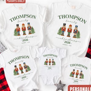 May include: Four white short-sleeved shirts and one white baby bodysuit featuring a graphic of three nutcrackers, green pine trees, and the text "THOMPSON family christmas 2026 ASPEN - CO". The shirts and bodysuit are arranged together.