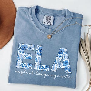 May include: A light blue t-shirt with the letters "ELA" in a floral pattern. The words "english language arts" are written below the letters in a script font. A gold necklace and a brown hat are also in the image.