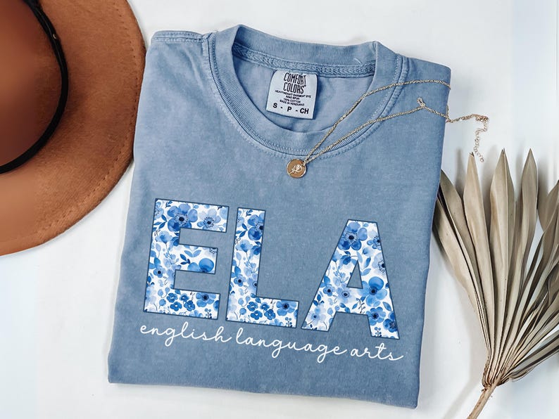 May include: A light blue t-shirt with the letters "ELA" in a floral pattern. The words "english language arts" are written below the letters in a script font. A gold necklace and a brown hat are also in the image.