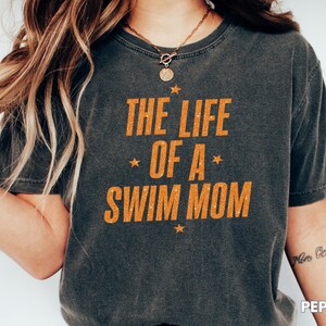 May include: Dark gray t-shirt with the text "THE LIFE OF A SWIM MOM" in orange. The text is accented with stars. The shirt is a casual style.