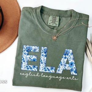 May include: Moss green t-shirt with the letters "ELA" in a blue floral pattern. The words "english language arts" are printed below the letters in a cursive font. A gold necklace is draped across the shirt.